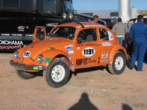 1990 Ford Bronco - orange VW Beetle-based Baja Bug, marked 1191 and lettered for Pump Gas Racing, sits on dirt beside haulers at a desert race.