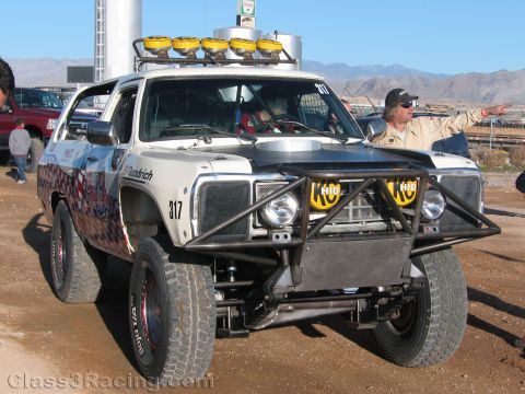 1990 Ford Bronco - silver off-road race SUV numbered 317 with a roof-mounted light bar and tubular front bumper stands on dirt with desert mountains behind.