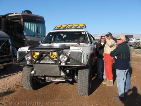 1990 Ford Bronco - Spectators talk beside the front of a silver off-road race SUV numbered 317 fitted with yellow driving lights, parked near a motorhome.