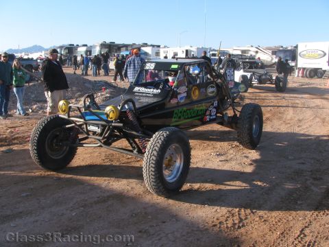 1990 Ford Bronco - black tube-frame off-road race buggy with BFGoodrich graphics sits in a busy dirt pit area surrounded by spectators and trailers.