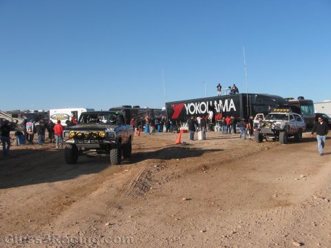 1990 Ford Bronco - Off-road race trucks gather on a dirt lot beside a black Yokohama-branded semi-trailer as crowds of people mill around under a blue sky.