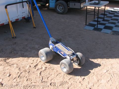 1990 Ford Bronco - chrome floor jack with a blue handle and four small wheels rests on sandy ground in a race pit beside a checkered mat.