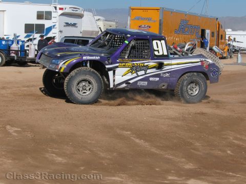 1990 Ford Bronco - purple and white off-road race truck numbered 91 kicks up a cloud of dirt as it corners past trailers at a desert race.
