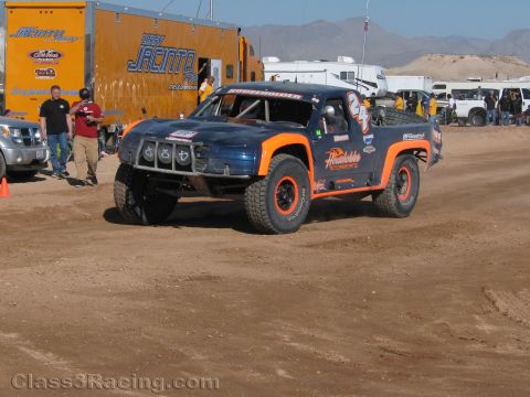 1990 Ford Bronco - blue and orange off-road race truck kicks up dirt on a desert course as a man walks past parked trailers and trucks behind.