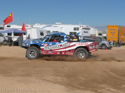 1990 Ford Bronco - off-road race truck numbered 71 in stars-and-stripes livery with General Tire and Lucas Oil branding throws dirt past RVs at a desert race.