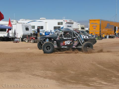 1990 Ford Bronco - black off-road race truck numbered 22 with K&N graphics kicks up dirt as it races across a desert course lined with trailers.