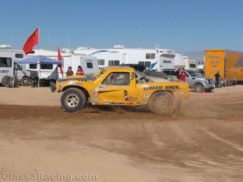 1990 Ford Bronco - yellow off-road race truck lettered Greer Bros and Toyo Tires kicks up dirt while cornering past RVs and a Chinese flag at a desert race.