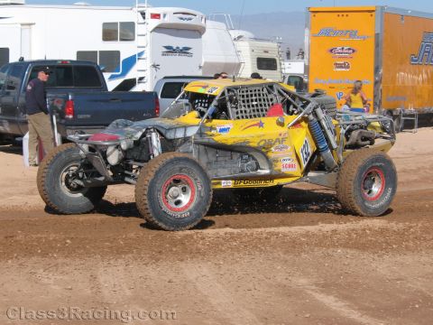 1990 Ford Bronco - yellow open tube-frame off-road race buggy on BFGoodrich tires sits on dirt among parked pickups and trailers at a desert race.