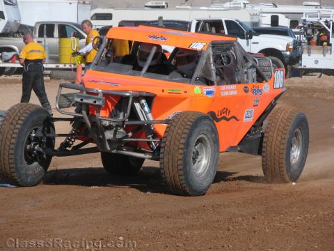 1990 Ford Bronco - orange single-seat off-road race buggy numbered 1106 sits on dirt as crew in yellow shirts work nearby at a desert race.
