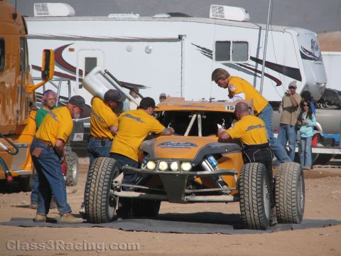 1990 Ford Bronco - pit crew in yellow shirts pushes a gray off-road race buggy across dirt in front of large motorhomes at a desert event.