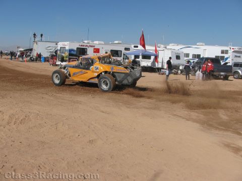 1990 Ford Bronco - yellow off-road race buggy numbered 154 throws up dirt while racing along a desert course lined with trailers and spectators.