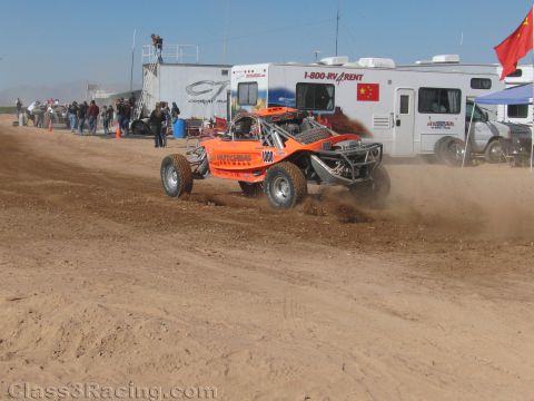 1990 Ford Bronco - orange off-road race buggy kicks up dirt while powering along a banked desert course in front of parked RVs and spectators.
