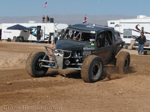 1990 Ford Bronco - black off-road race buggy numbered 116 with BFGoodrich graphics throws dirt while cornering at a desert race near RVs and flags.