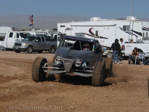 1990 Ford Bronco - dark blue tube-frame off-road race buggy throws dirt while cornering on a desert course in front of parked pickups and RVs.