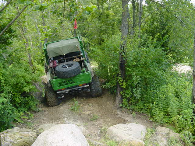 Green off-road vehicle with spare tire mounted on the back, driving on a dirt trail surrounded by trees