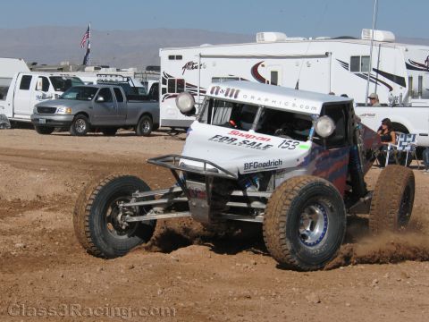 1990 Ford Bronco - white off-road race buggy numbered 153 and marked For Sale throws dirt while cornering at a desert race in front of RVs and pickups.