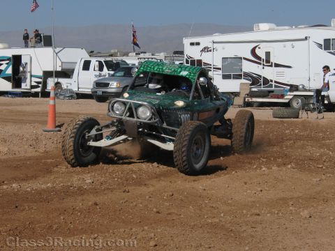 1990 Ford Bronco - green off-road race buggy kicks up dirt while cornering on a desert course in front of pickups, RVs and a flag.