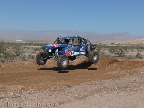 1990 Ford Bronco - blue off-road race buggy lettered Malicious catches air off a dirt berm with hazy desert mountains in the background.