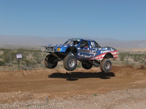 1990 Ford Bronco - blue stars-and-stripes off-road race truck numbered 41 with General Tire branding jumps a dirt berm against a desert backdrop.