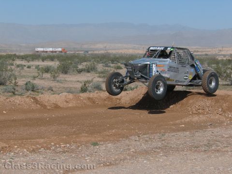 1990 Ford Bronco - silver off-road race buggy catches air off a dirt berm on an open desert course with mountains in the distance.