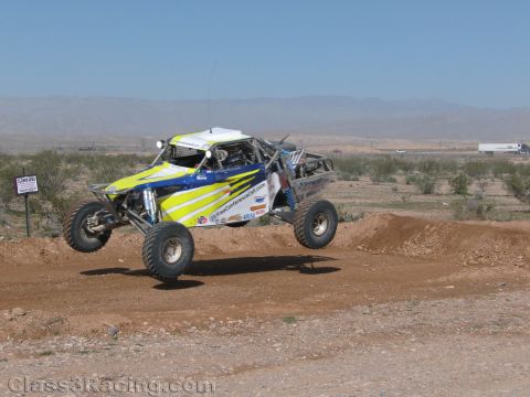 1990 Ford Bronco - yellow and blue off-road race buggy launches off a dirt berm on an open desert course with hazy mountains behind.