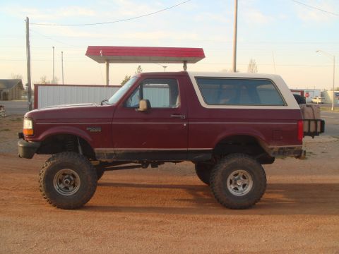 1992 Ford Bronco - Driver-side profile of a lifted burgundy Ford Bronco with a white removable hardtop, black flat front bumper and Bronco fender badge, stand…