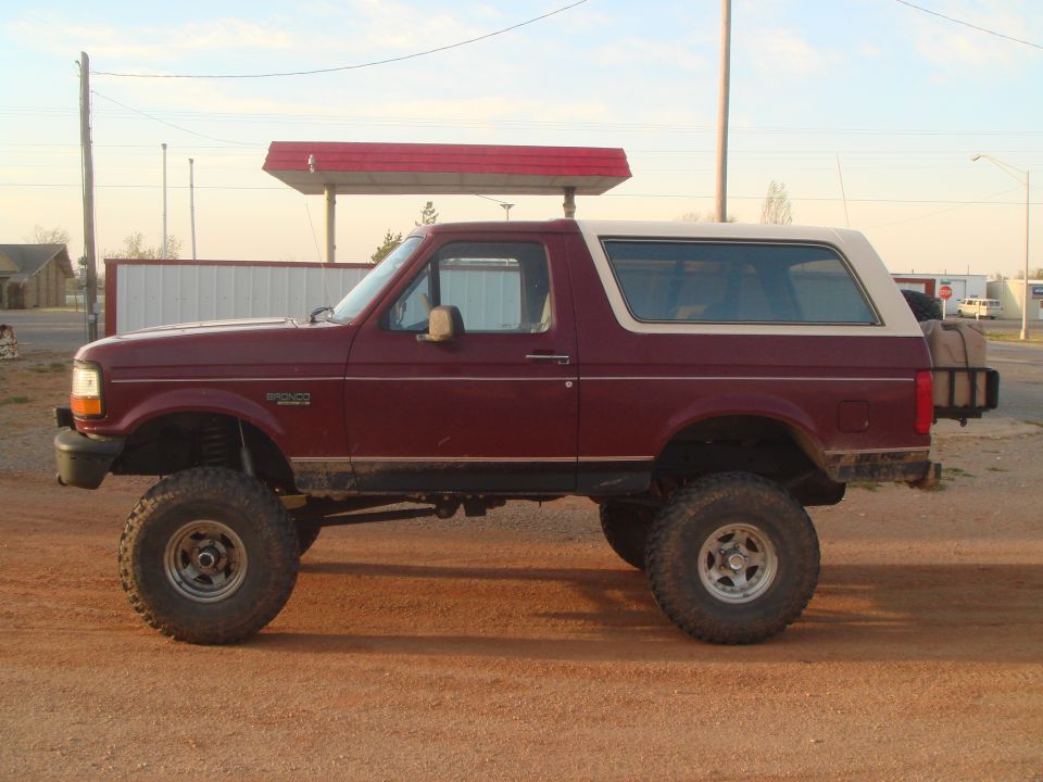 Side profile of lifted burgundy Ford Bronco with white top on mud tires, red dirt lot