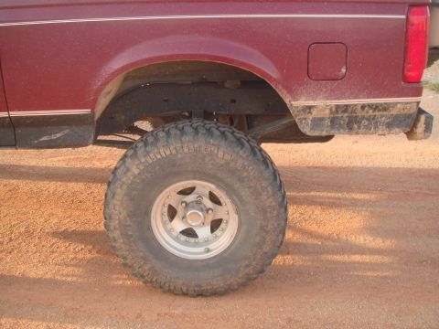 1992 Ford Bronco - Close side view of the rear wheel well on a lifted burgundy Ford, showing a big mud-terrain tire on a polished slotted aluminum wheel with…