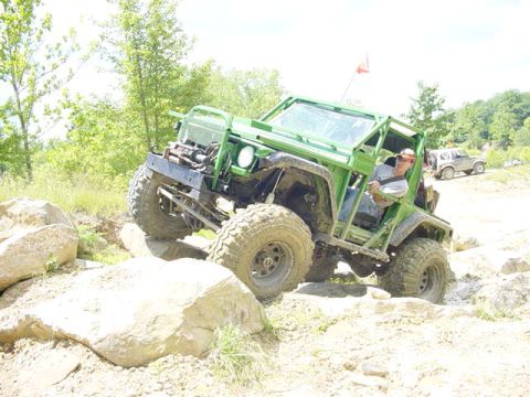 1992 Ford Bronco - A heavily modified green off-road vehicle navigates a rocky terrain, with another vehicle visible in the distance.