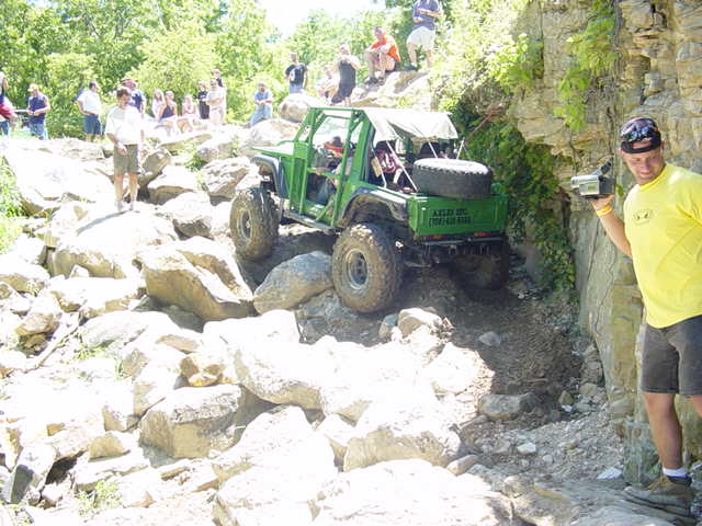 Green off-road vehicle with a spare tire on the back climbs a rocky incline, with spectators watching