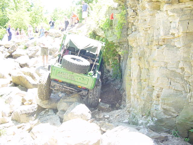 green off-road vehicle with spare tire on back climbing over large rocks near a cliff face, people watching