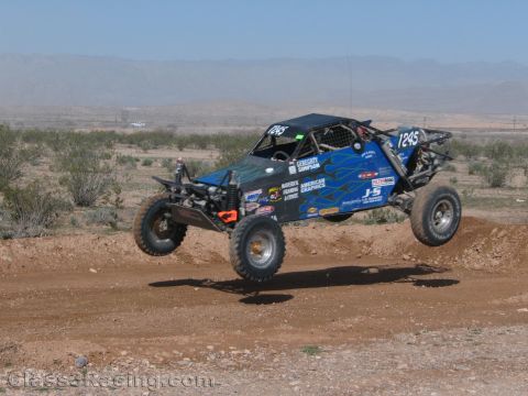 1990 Ford Bronco - blue off-road race buggy numbered 1245 catches air off a dirt berm on an open desert course with distant mountains.