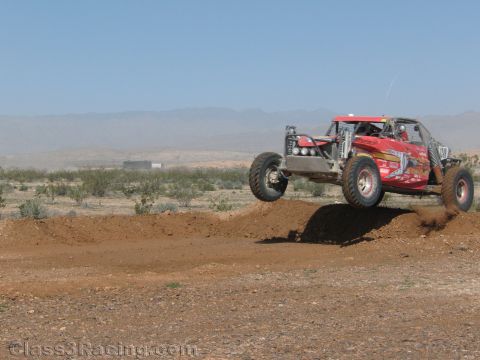 1990 Ford Bronco - red single-seat off-road race buggy launches off a dirt berm with desert scrub and distant mountains behind.