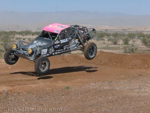 1990 Ford Bronco - off-road race buggy numbered 1299 with a pink roof panel sails over a dirt berm in the desert.