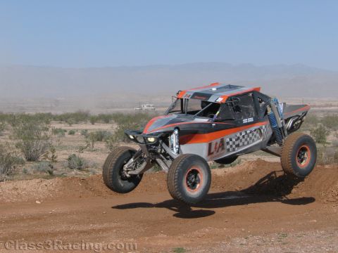 1990 Ford Bronco - orange-and-gray off-road race buggy with a checkered side panel jumps over dirt with desert terrain behind.