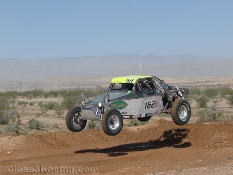 1990 Ford Bronco - gray off-road race buggy numbered 1622 with a yellow-green roof catches air over a dirt berm in the desert.