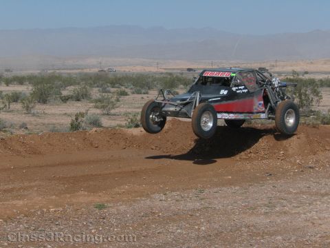 1990 Ford Bronco - dark off-road race buggy with red accents, numbered 54, jumps a dirt berm across an open desert course.