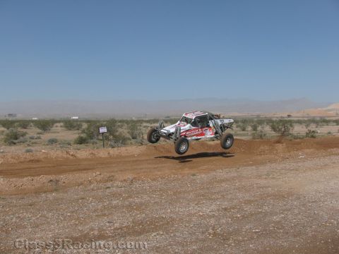1990 Ford Bronco - white-and-red off-road race buggy catches air over a dirt jump, seen at a distance across the desert.