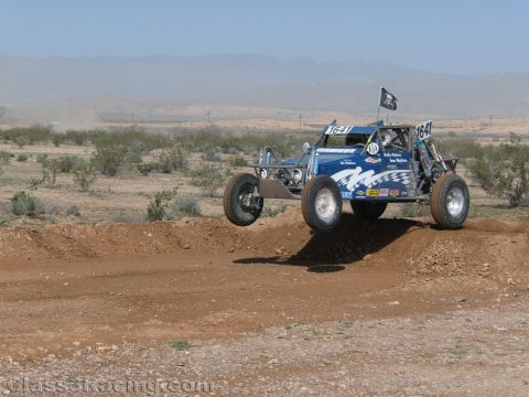1990 Ford Bronco - blue off-road race buggy numbered 1647 flying a Jolly Roger flag jumps a dirt berm in the desert.