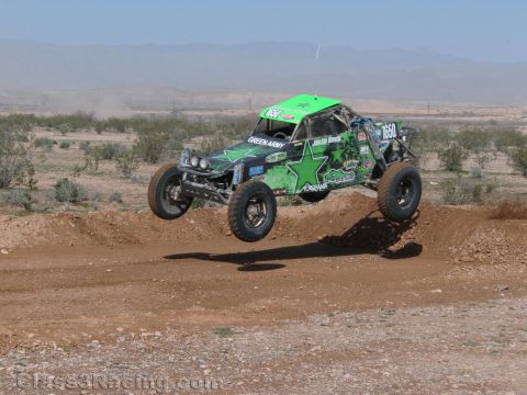 1990 Ford Bronco - green off-road race buggy with star graphics and Green Army livery catches air over a dirt berm in the desert.