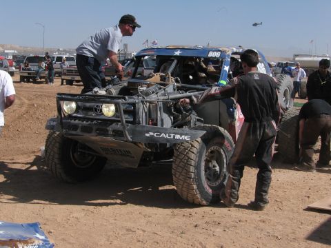 1990 Ford Bronco - crew works on a stripped off-road race truck numbered 806 in a busy, dusty desert pit area with a helicopter overhead.
