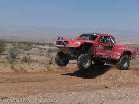 1990 Ford Bronco - red off-road race truck with roof lights catches air over a dirt berm in an open desert race setting.