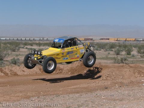 1990 Ford Bronco - yellow off-road race buggy leaps a dirt berm in the desert with a freight train visible in the background.