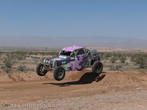1990 Ford Bronco - purple off-road race buggy with star graphics catches air over a dirt berm on a desert race course.