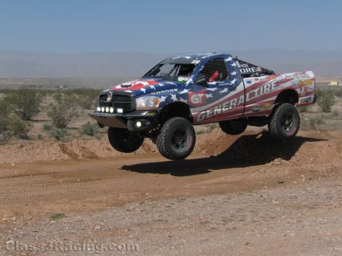 1990 Ford Bronco - Dodge Ram off-road race truck in an American-flag wrap with General Tire livery launches off a dirt jump in the desert.