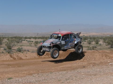 1990 Ford Bronco - Volkswagen Beetle-based Baja race bug numbered 552 with a red roof jumps a dirt berm in the desert.