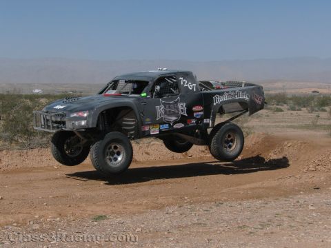 1990 Ford Bronco - gray off-road race truck numbered 1287 catches air over a dirt course in an open desert setting.
