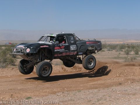1990 Ford Bronco - black off-road race truck numbered 713 with motorsports livery leaps a dirt berm across a desert course.