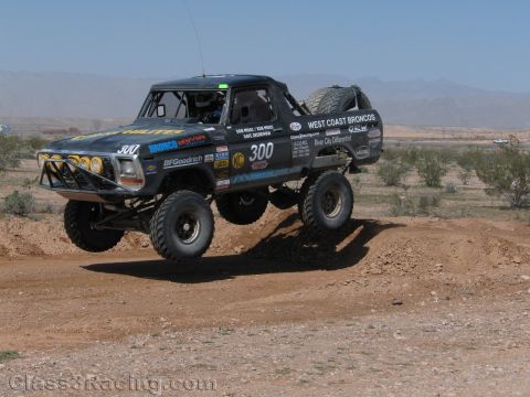 1990 Ford Bronco - vintage Ford Bronco off-road race truck numbered 300 with West Coast Broncos livery jumps a dirt berm in the desert.