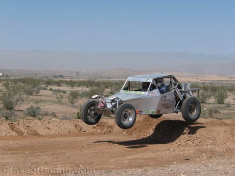 1990 Ford Bronco - silver off-road race buggy catches air over a dirt berm on an open desert race course.